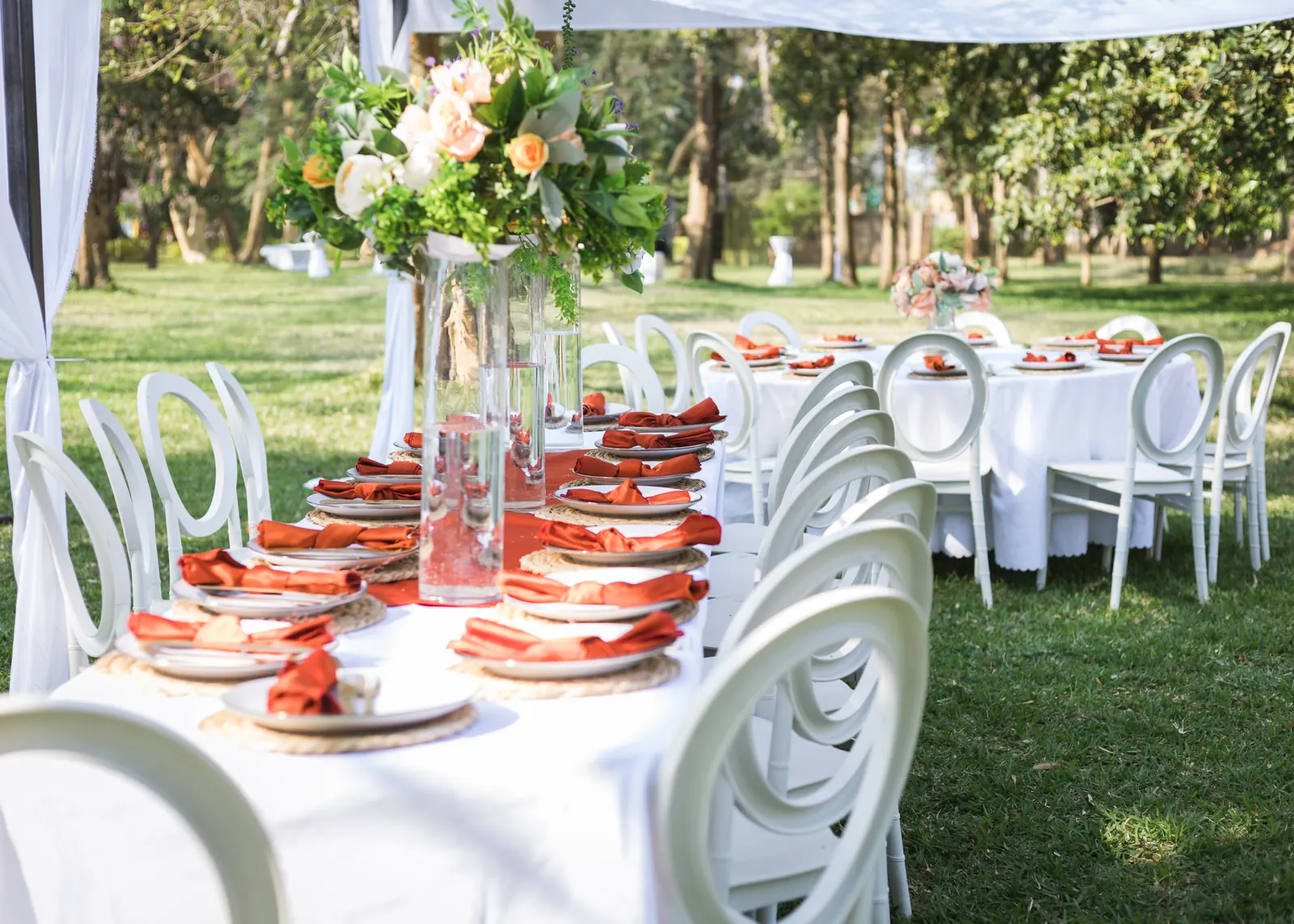 Outdoor wedding reception tables set with white chairs, peach flowers and orange napkins, ready for guests